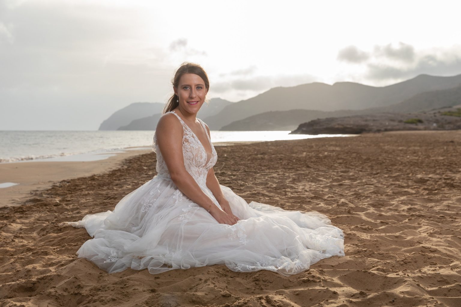 Novia sentada en la arena con la costa al fondo